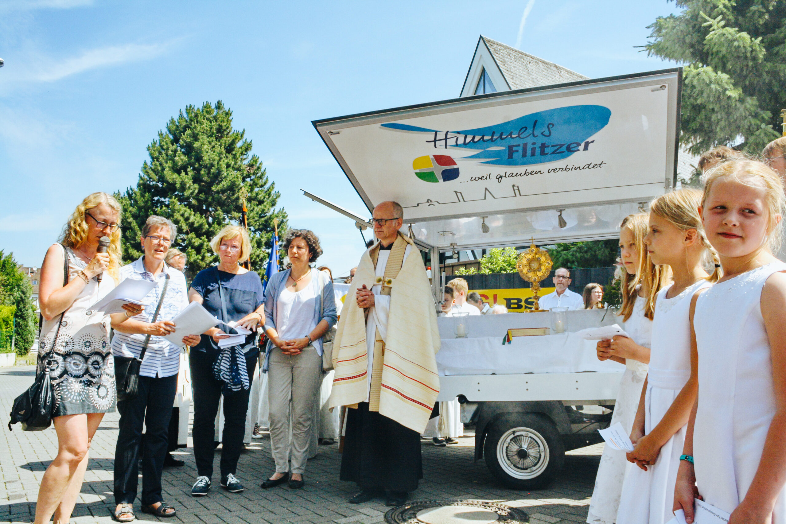 Kommunionkinder, Priester und Gemeindeangehörige stehen zu Fronleichnam vor dem Kirchenmobil HimmelsFlitzer.